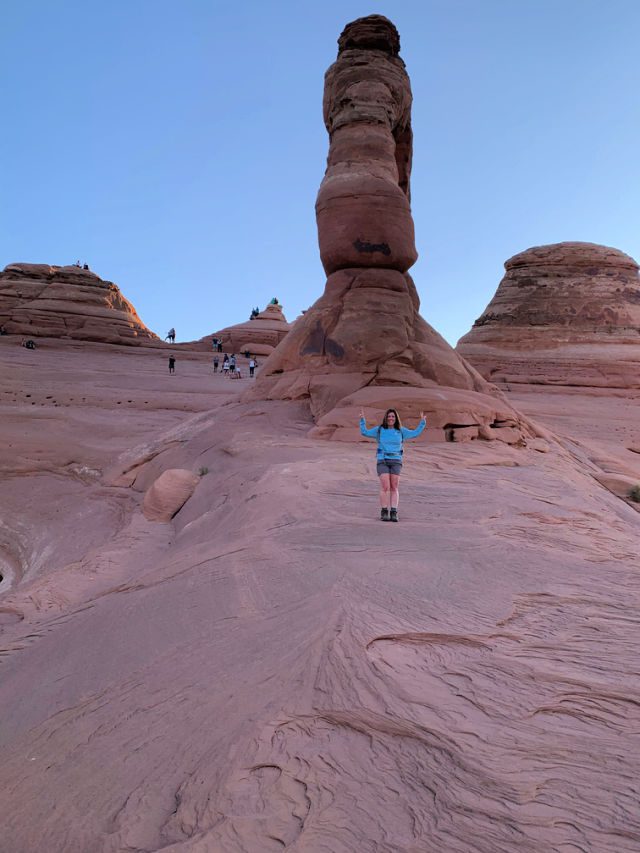 Lori standing to the side of Delicate Arch with viewers on The opposite side enjoying the views of the arch. 