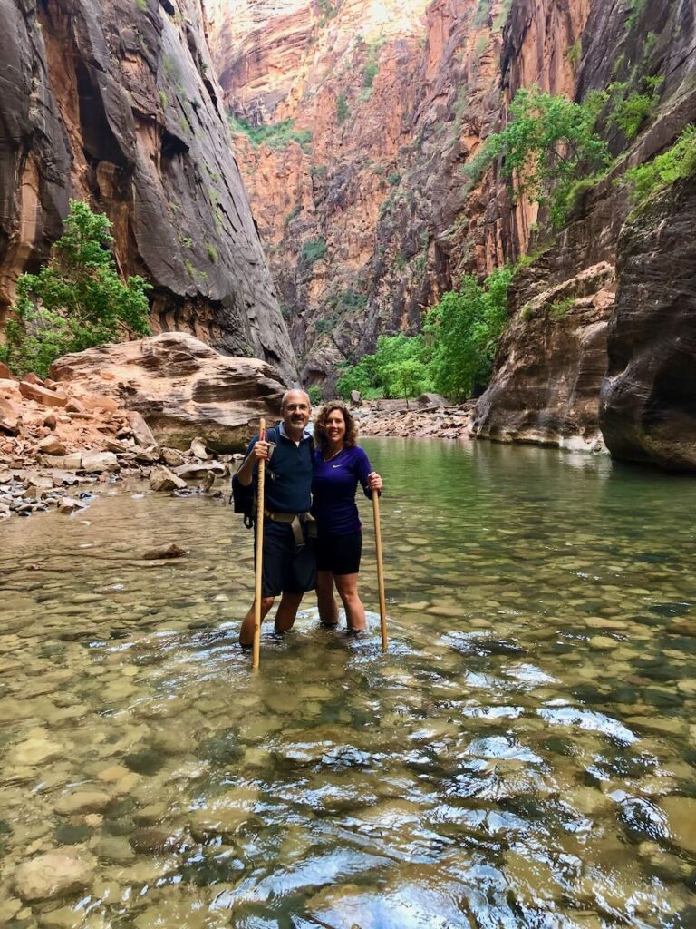 Lori and Steve are standing in the river and posing for a photo at the start of The Narrows hike